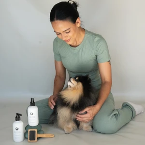 Woman in sage green ribbed top and trousers kneeling on a seamless grey studio floor, petting a fluffy Pomeranian dog.