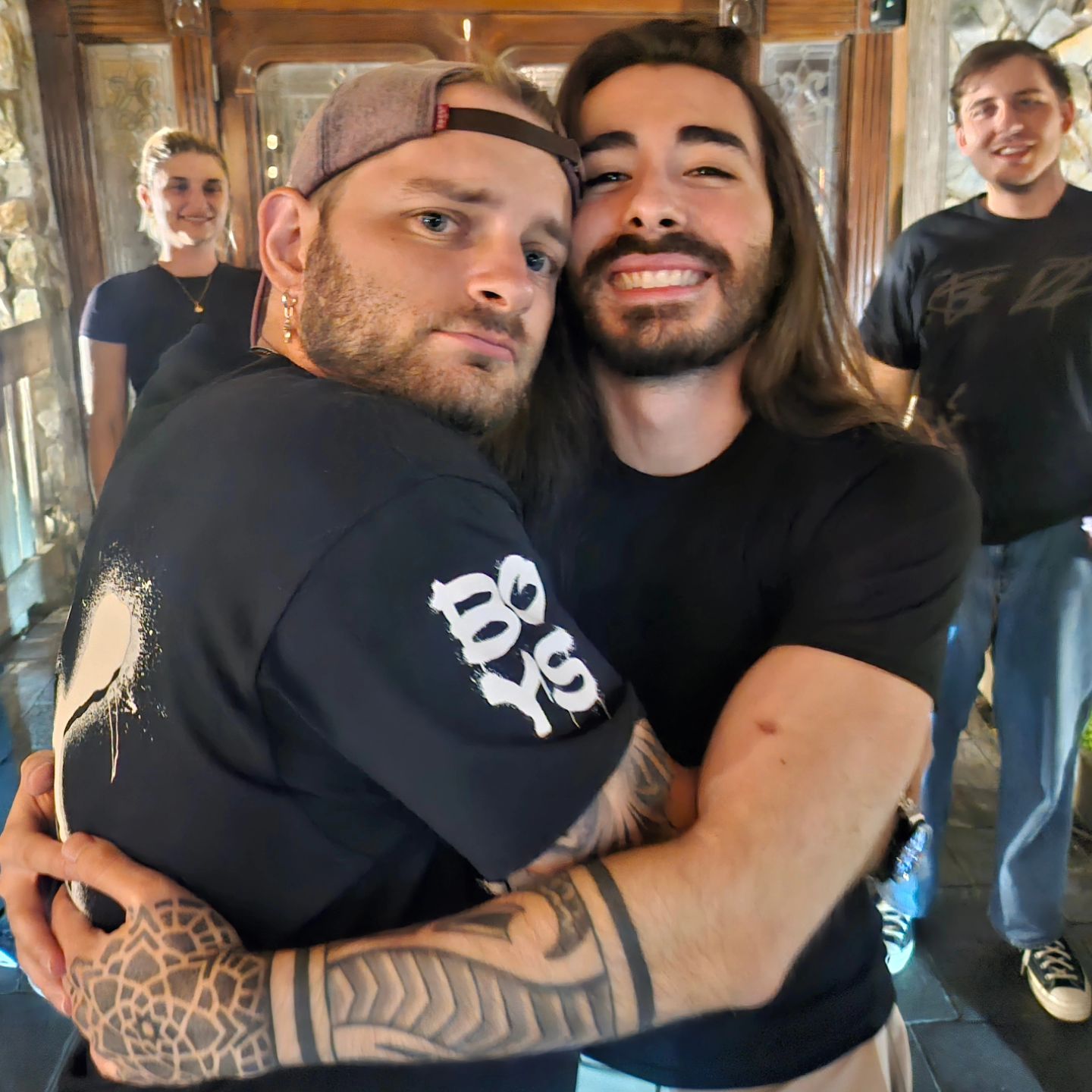 Two adult men embrace indoors, both wearing dark shirts and casual attire. Additional people are partially visible in the background.