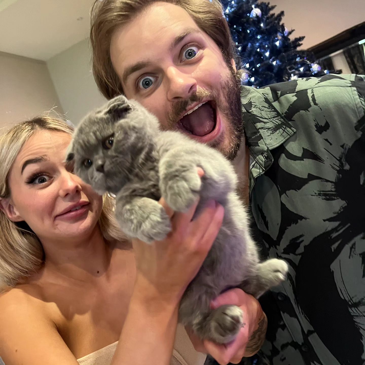 A joyful couple poses indoors, holding a grey kitten; a Christmas tree is visible in the background, adding a festive touch to the scene.