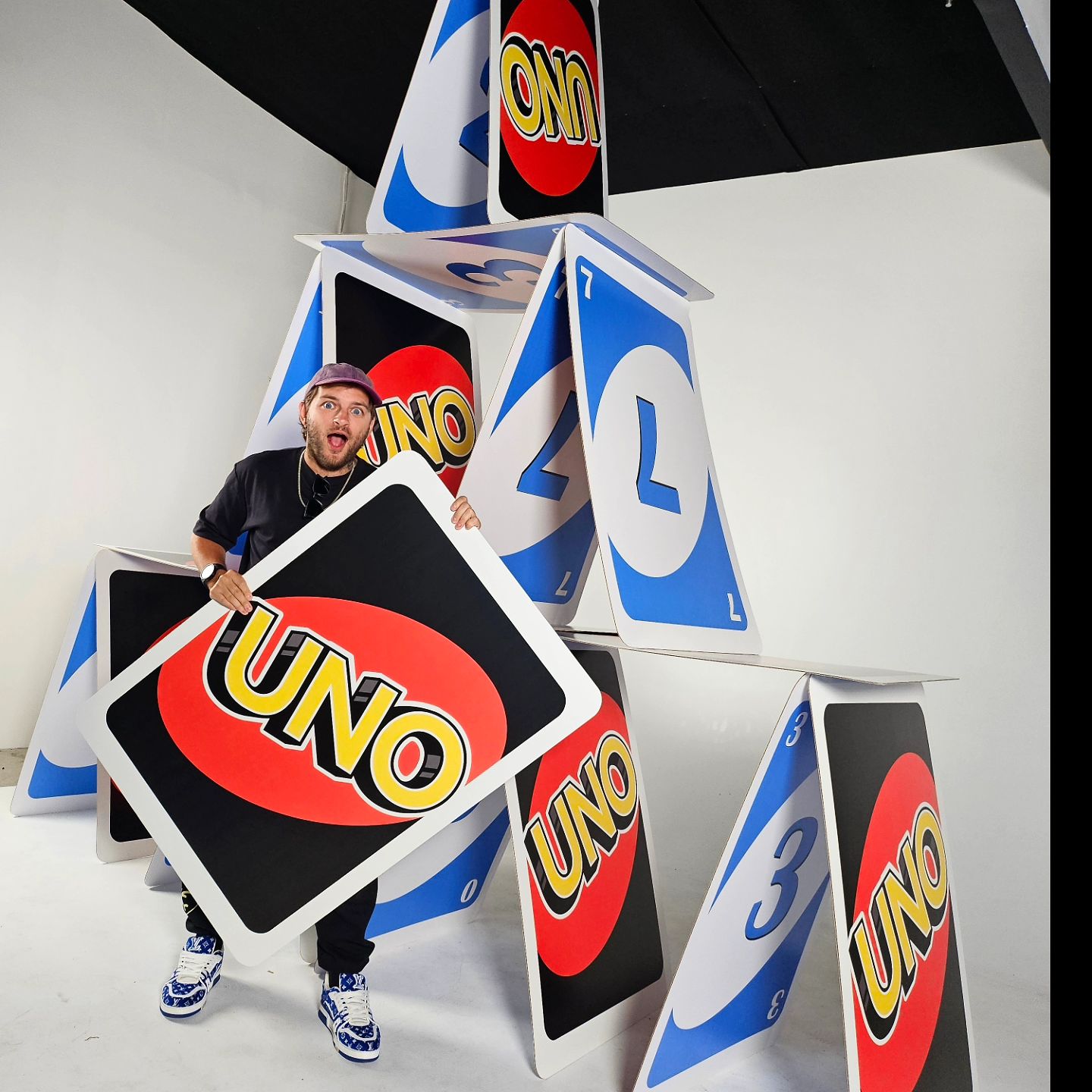 Young man poses playfully with oversized UNO cards in a bright studio setting, forming a whimsical, architectural structure with the cards.