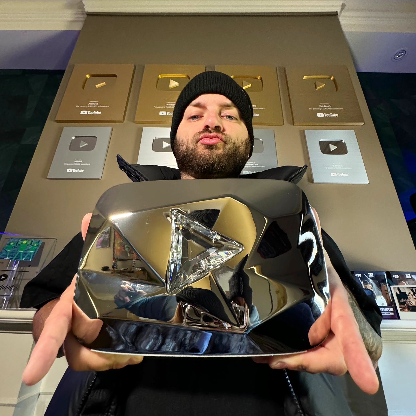Adult male holding a YouTube Diamond Play Button award in an indoor setting with additional awards displayed on the wall behind him.