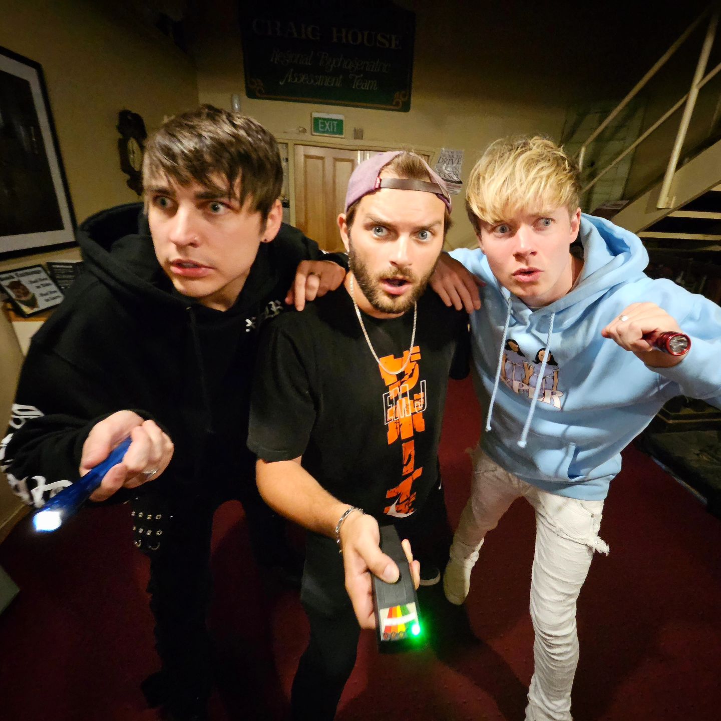 Three young adult men indoors point flashlights and a device with a light-up screen toward the camera, looking surprised in a dark room.