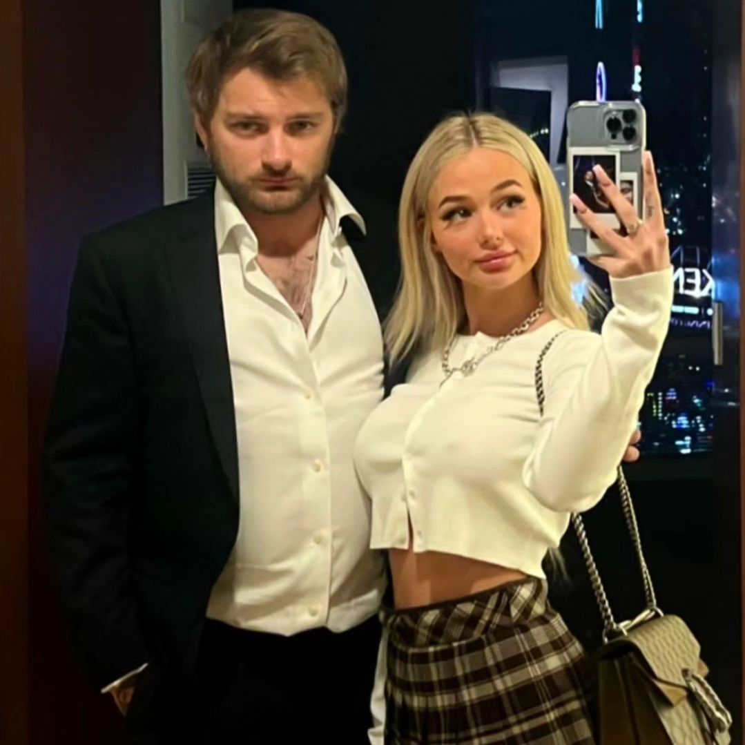 A blond woman in a skirt taking a selfie with a blond man in a suit in a hotel room at night, with city lights visible.