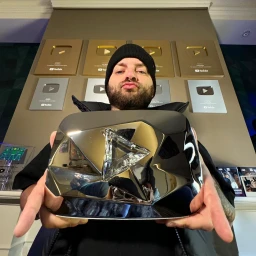 Content creator JoshDub holding a large reflective YouTube award plaque with a wall of subscriber buttons behind him