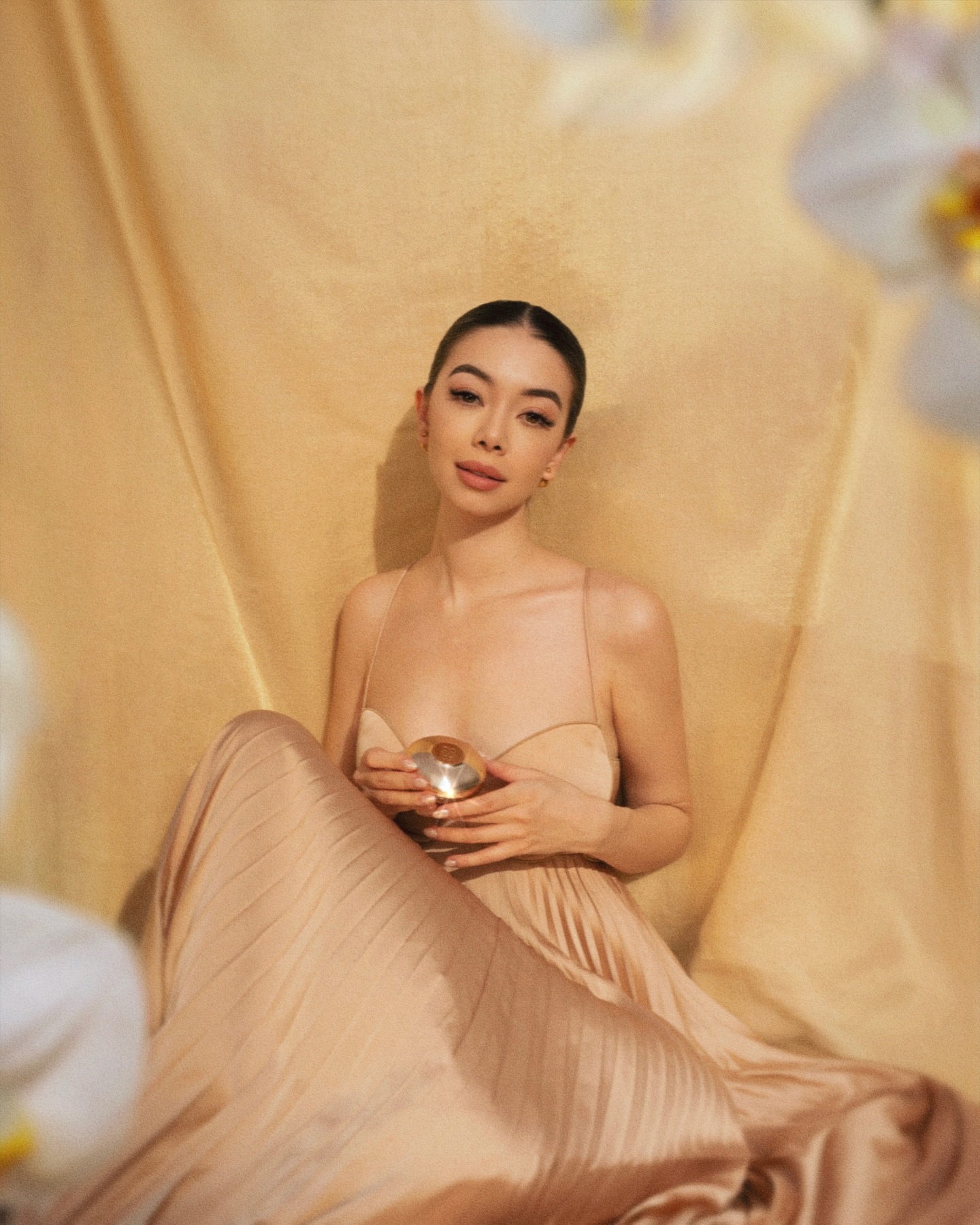 Elegant woman in a flowing champagne dress sits in front of a gold backdrop holding a small glass orb, surrounded by white orchids.