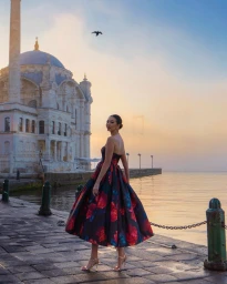 Woman in floral strapless dress standing on a stone pier by the water with a mosque in the background during sunset.
