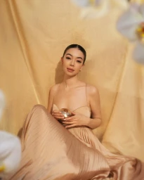 Woman in pleated champagne dress sitting against draped golden fabric backdrop holding small golden perfume bottle