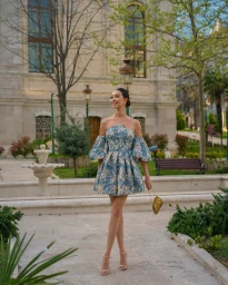 Woman wearing a blue patterned off-the-shoulder minidress standing in a stone courtyard with trees and a small fountain