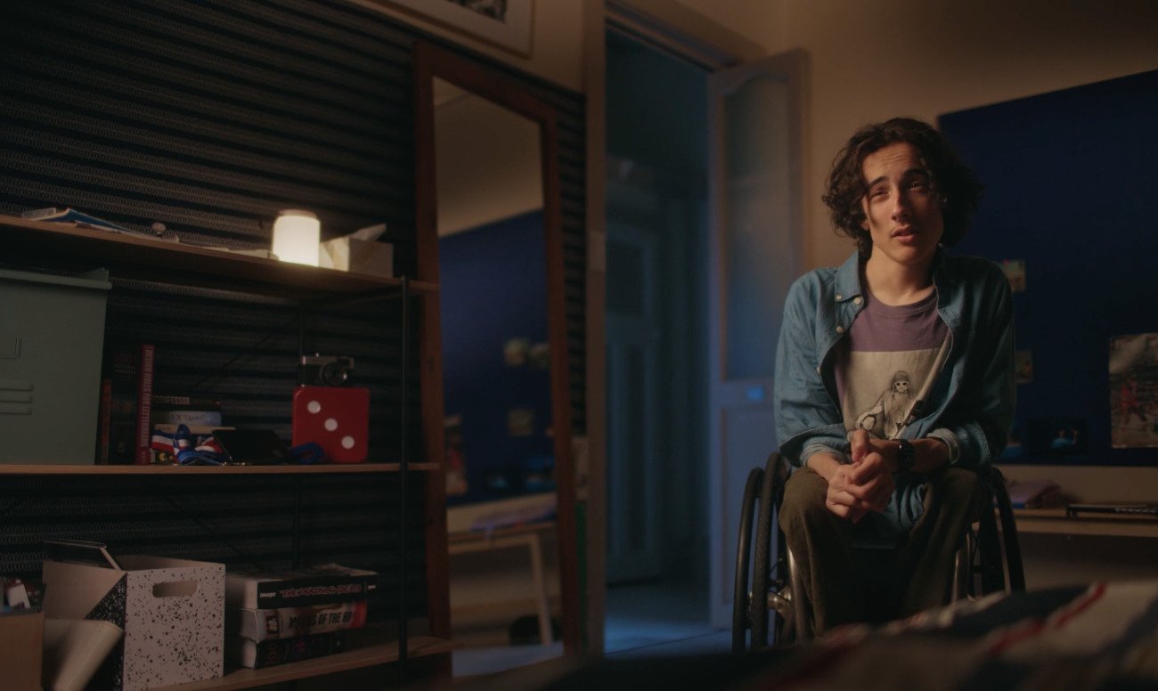 Young man in wheelchair sits in a room with shelves, desk, and decor; a mirror reflects the scene in dim, warm lighting.