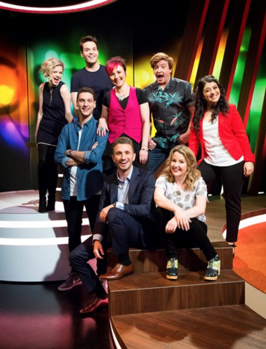Group photo of seven adults indoors in a studio with a colorful backdrop. They are standing and sitting on a wooden platform.