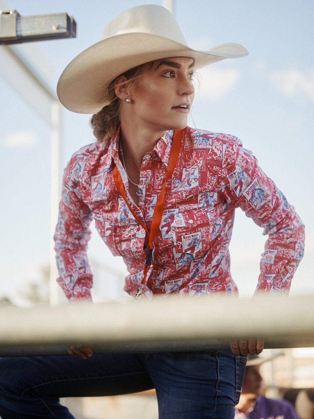 Portrait of a cowgirl wearing a cowboy hat and patterned shirt. The cowgirl is standing outdoors near a fence on a sunny day.