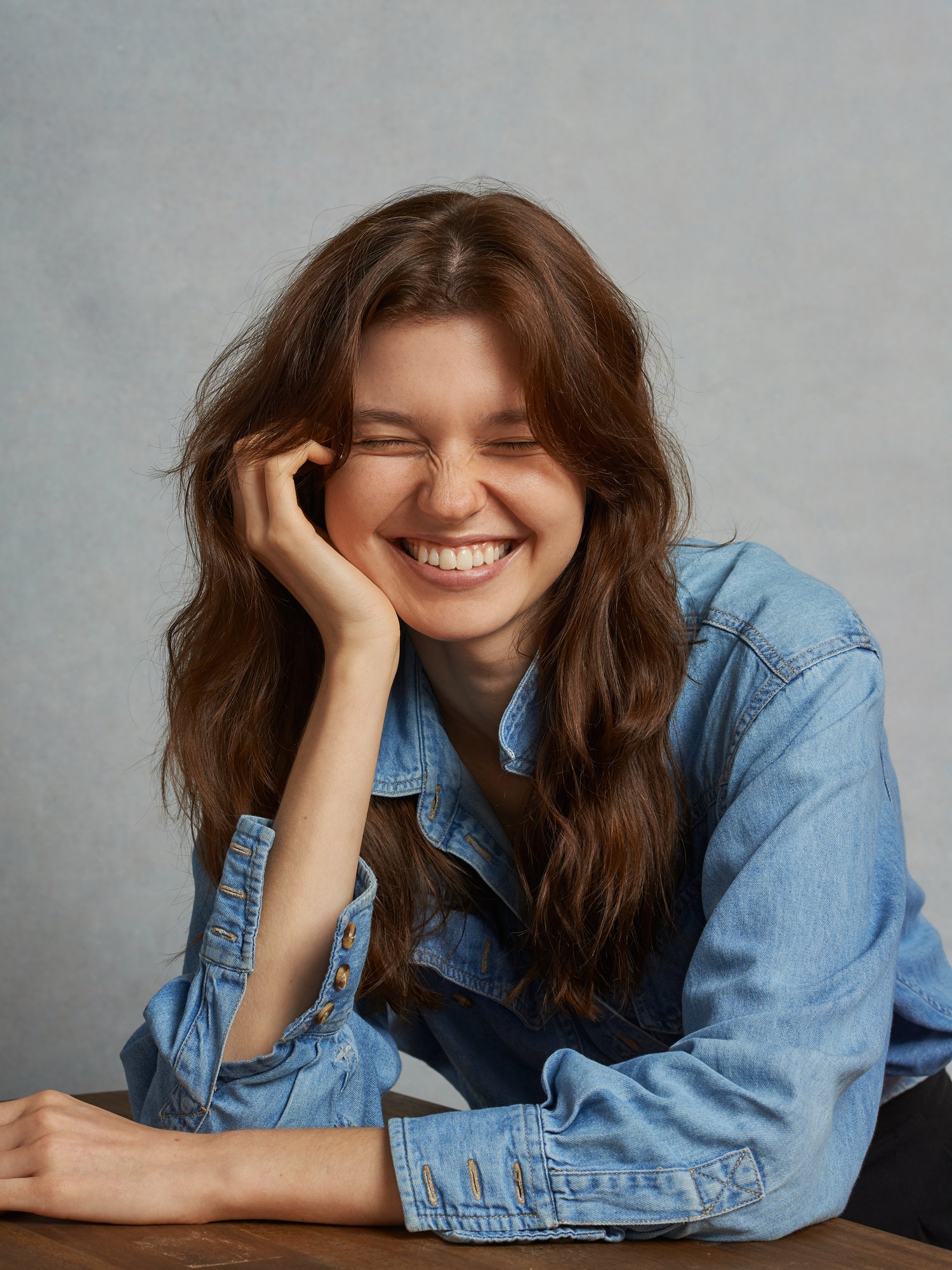 Smiling young woman with long brunette hair, wearing a blue denim shirt, rests her head on her hand in a close-up portrait.