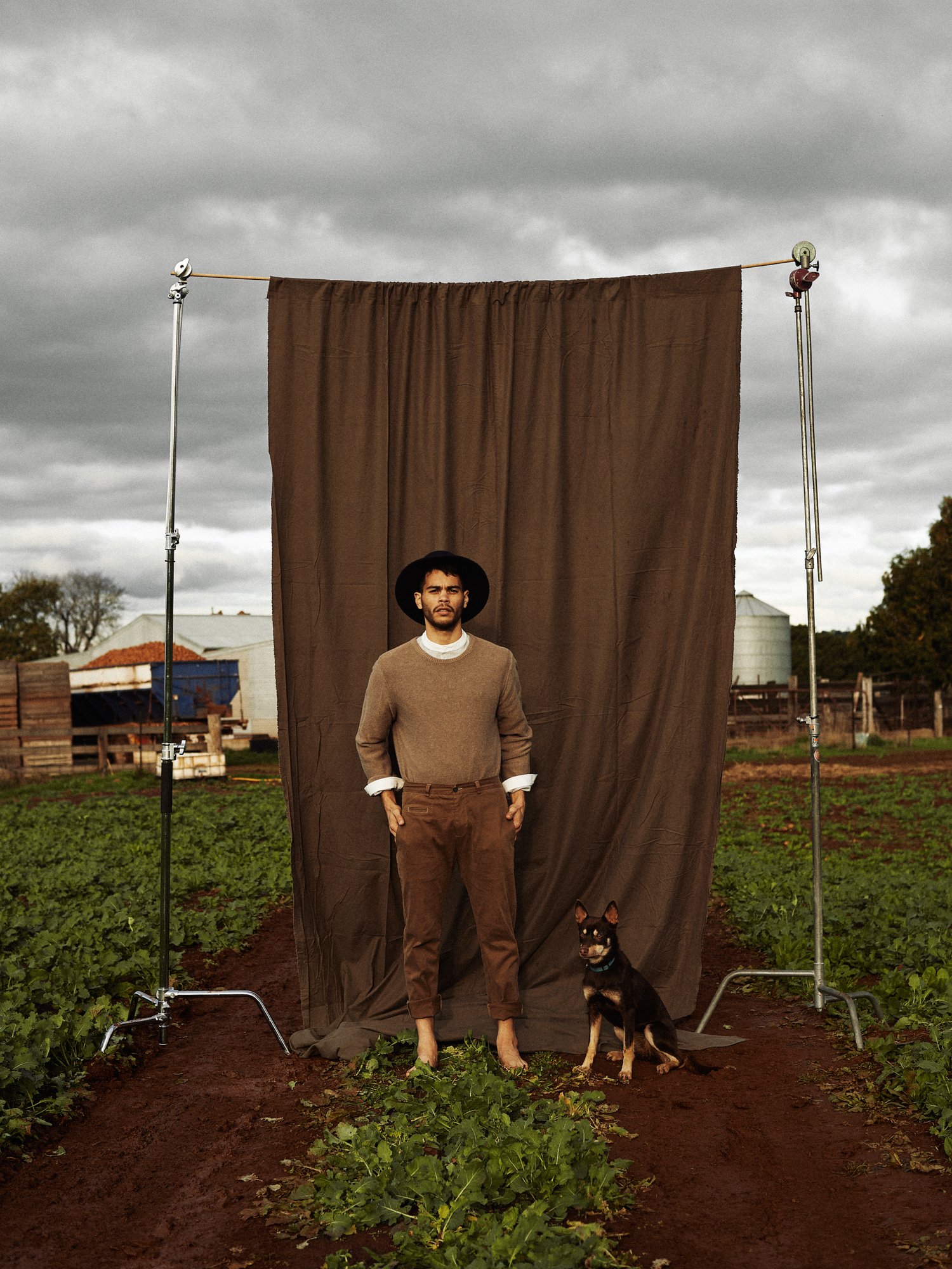 Barefoot man in brown clothing poses with dog near brown backdrop and crops in a rural setting under cloudy skies.