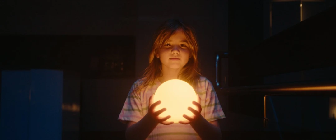 A young girl in a dimly lit room holds a glowing orb, casting a warm light on her face, creating a serene and magical atmosphere.