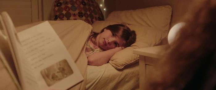 A young girl in bed looking toward an open storybook held by someone seated nearby in a softly lit bedroom