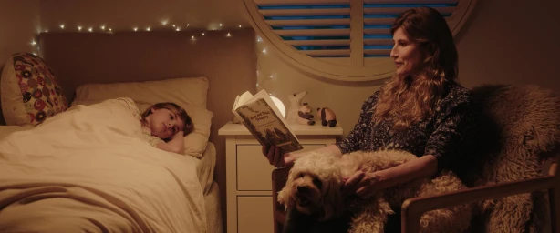 Woman reading a book to a child in bed while holding a small dog in a warm, dimly lit bedroom.