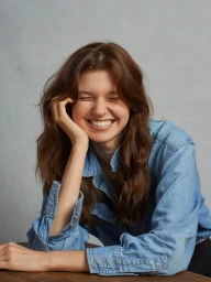 A smiling young woman with long brown hair wearing a denim shirt, resting her head on her hand against a grey backdrop.