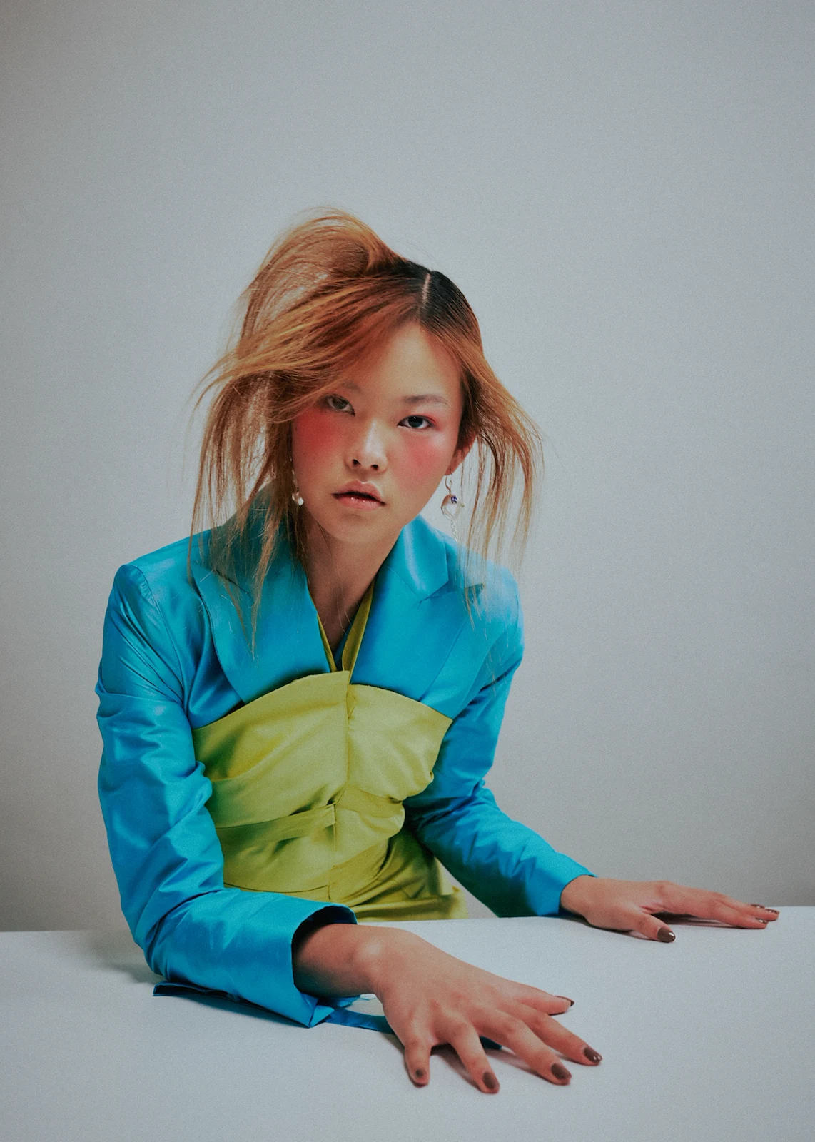 Asian female model poses with hands on table, wearing a teal jacket and light green top against a neutral studio backdrop.