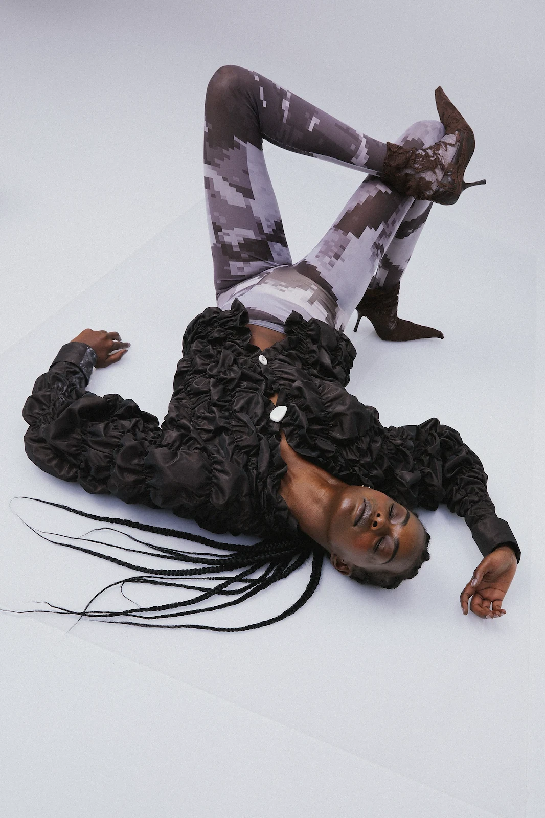 Fashion shot of a Black woman lying on her back wearing a ruffled black jacket, pixelated grey leggings, and brown lace-up heels.