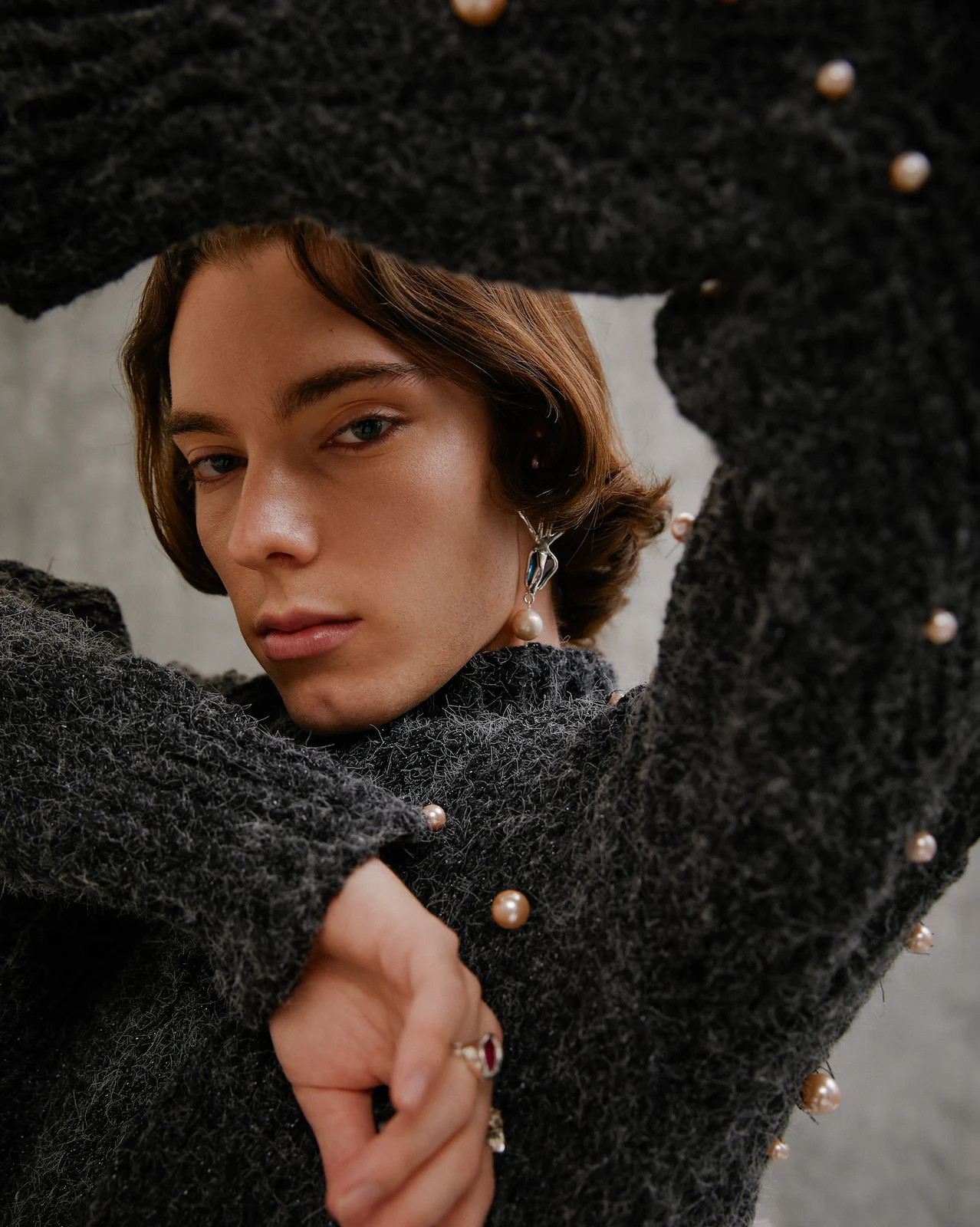 Portrait of a young adult with brown hair, wearing a pearl-studded gray sweater and pearl drop earrings, against a gray background.