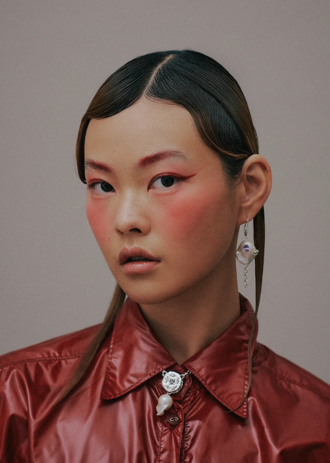 Portrait of a fashionable Asian woman with sleek hair and bold red makeup, wearing a red collared shirt and pearl earrings.