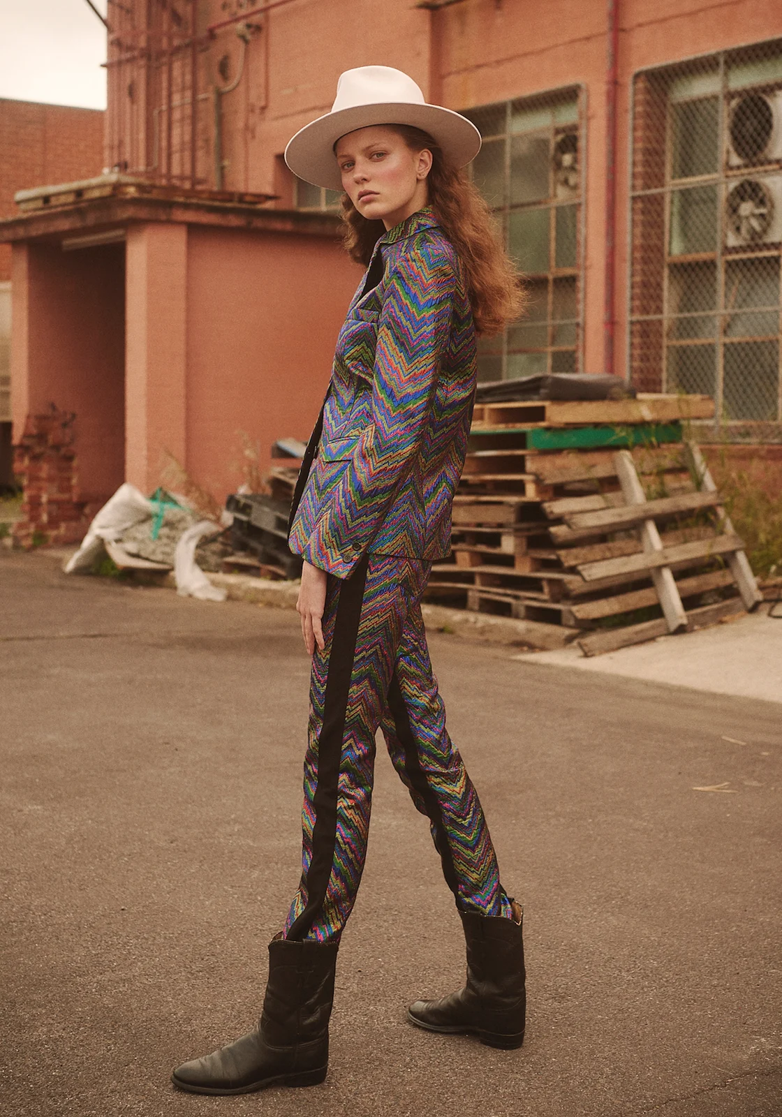 A female model in a patterned suit and cowboy boots stands on a paved road with a white hat, posing in front of a pink building.