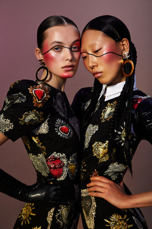 Two women pose in patterned dresses with exaggerated blush and black eyeliner. Fashion editorial shot against a neutral backdrop.