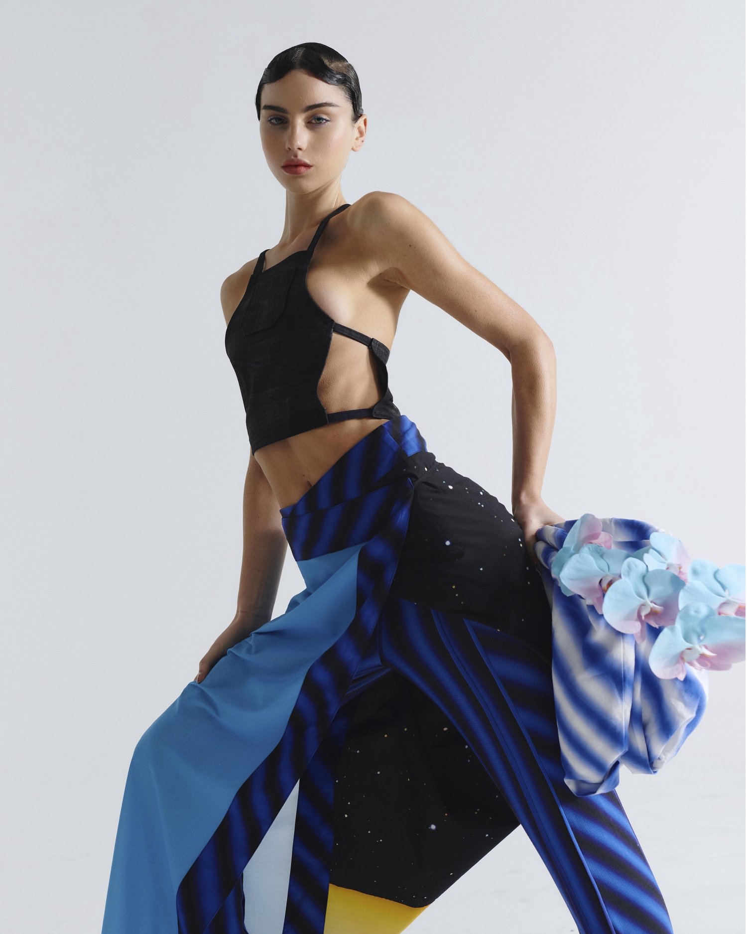 A woman with dark hair and red lipstick poses in a black crop top and colorfully patterned pants in a studio setting with neutral background.