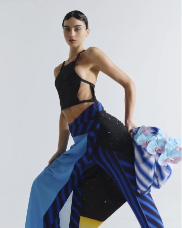 A woman with dark hair and red lipstick poses in a black crop top and colorfully patterned pants in a studio setting with neutral background.