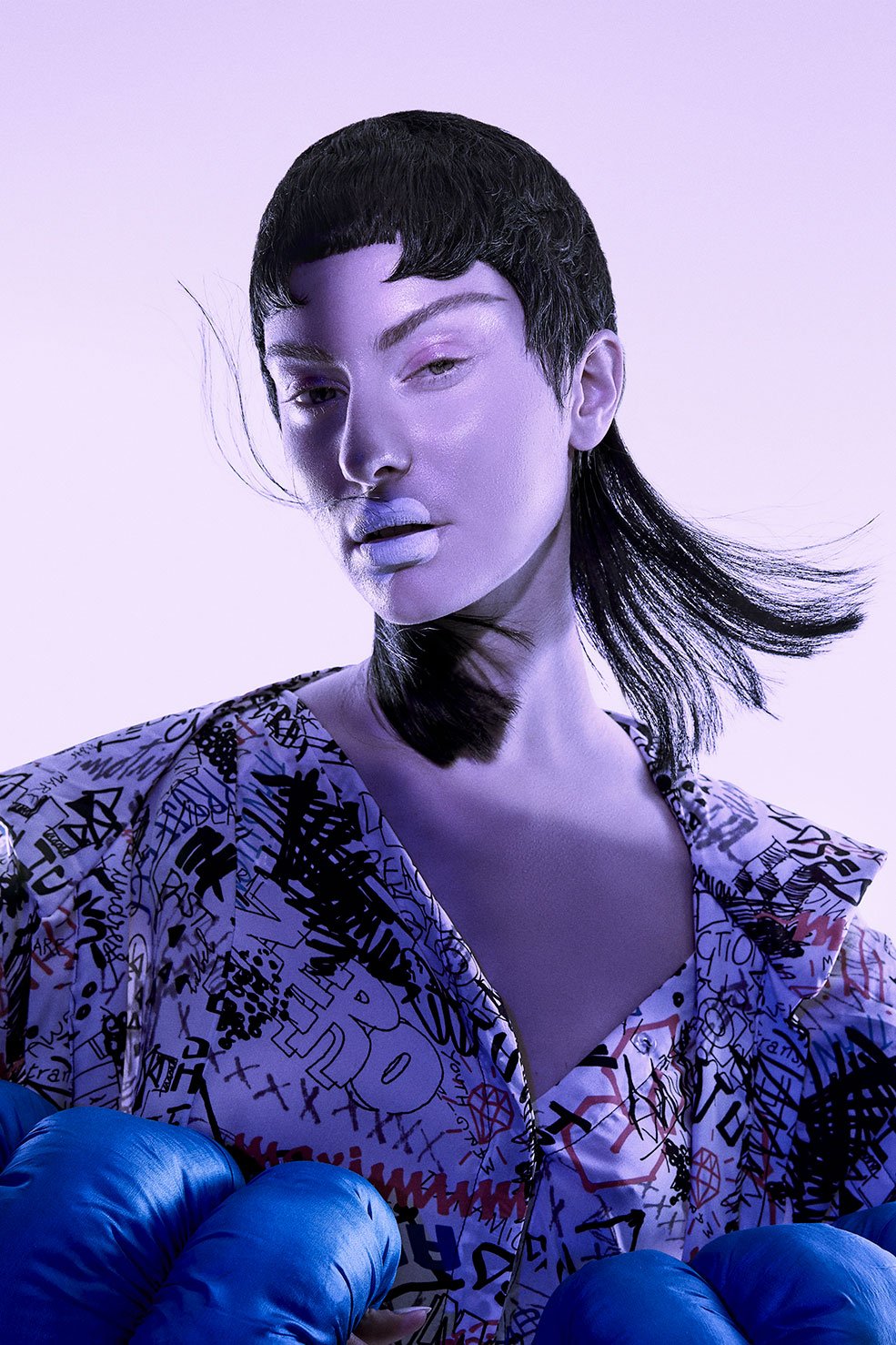 Portrait of an adult woman wearing a short black wig and a patterned blouse, showcasing vibrant blue boxing gloves, set against a lilac background.