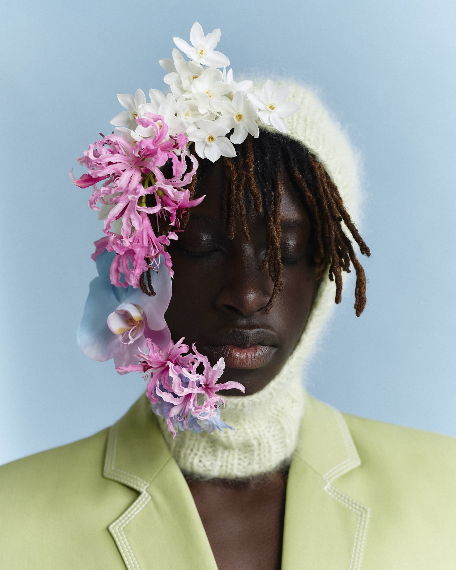 Dark-skinned model in light green blazer and knit hat decorated with pink and white flowers, posing against a light blue background.