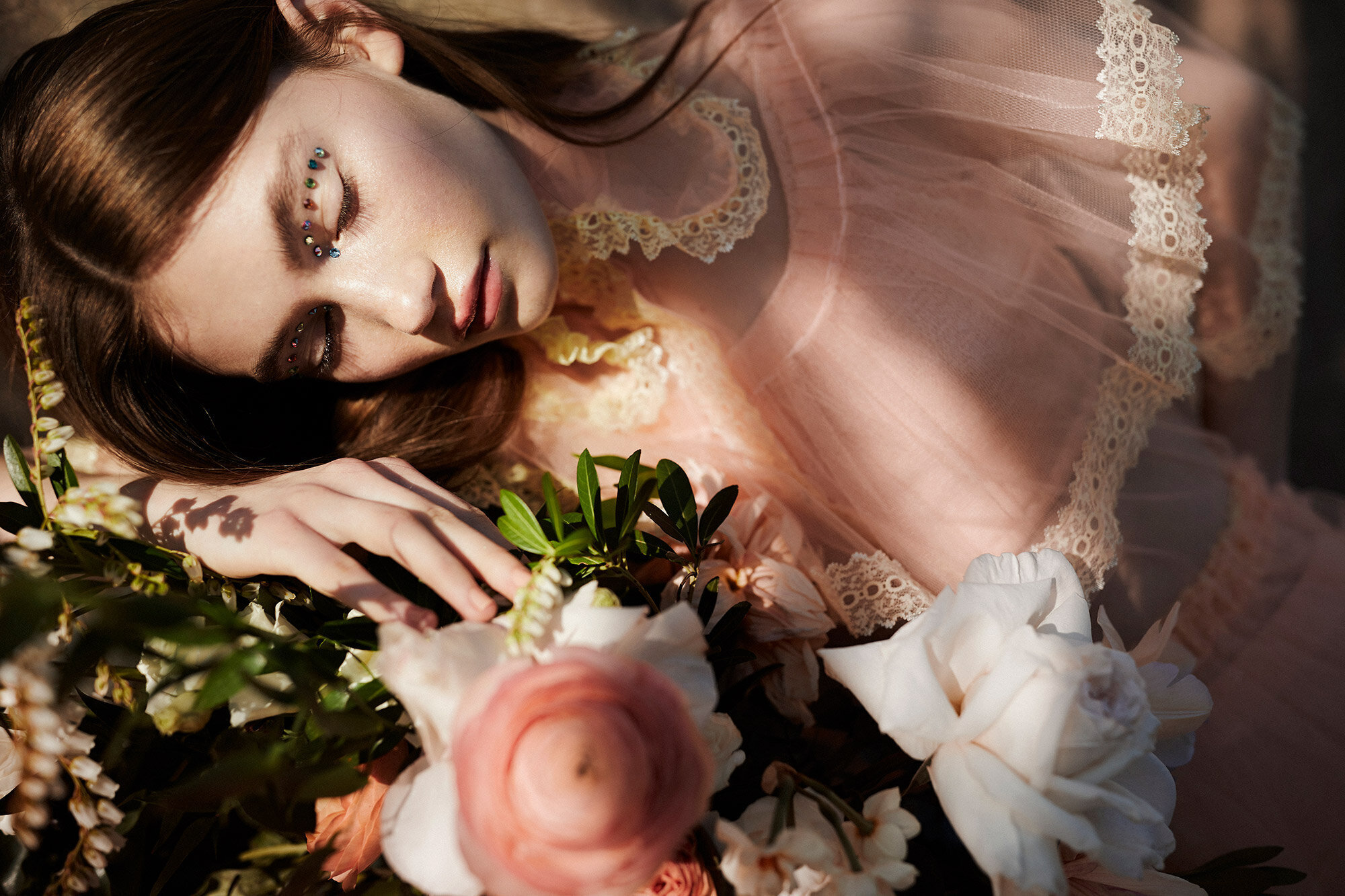 Woman in lace dress lies among white and pink flowers with colorful gems adorning her face in an outdoor setting with soft lighting.