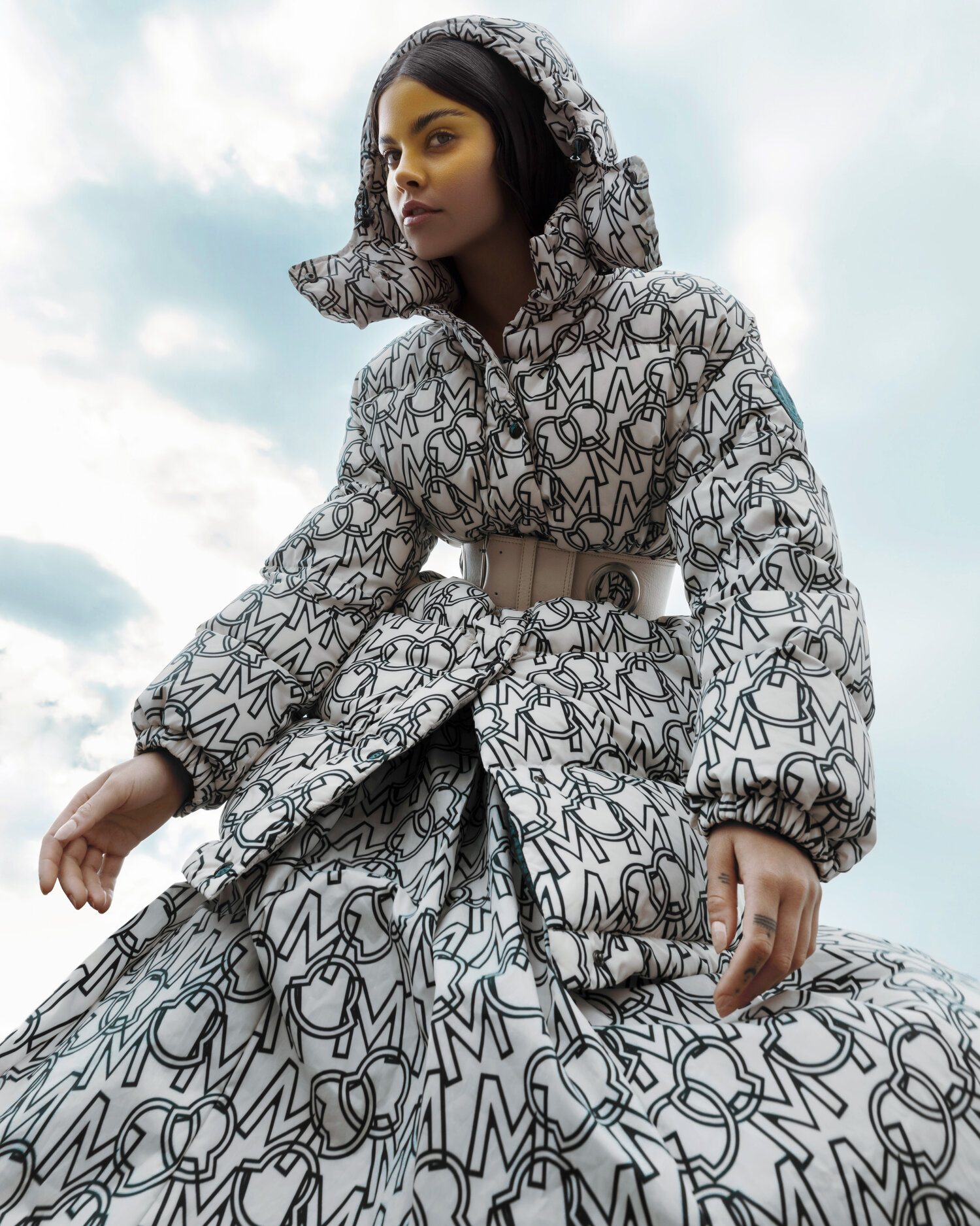 Fashion shot of a woman wearing a white puffy coat and skirt combo with a logo pattern. She is looking to the side against a cloudy sky.