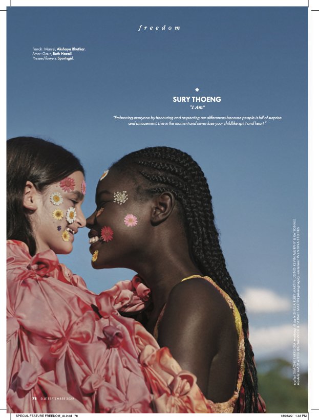 Editorial fashion shot of two women with floral decorations on their faces, embracing against a clear blue sky backdrop, soft lighting.