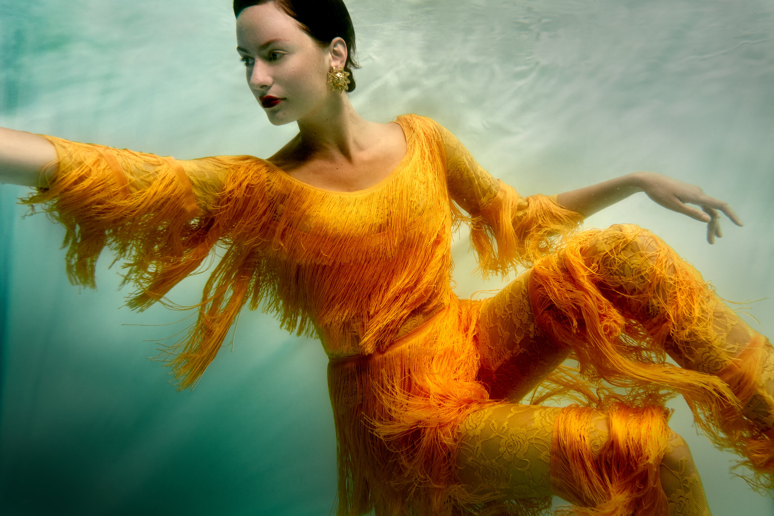 Elegant woman in a vibrant orange fringed dress poses gracefully underwater, showcasing a blend of fashion and ethereal beauty.