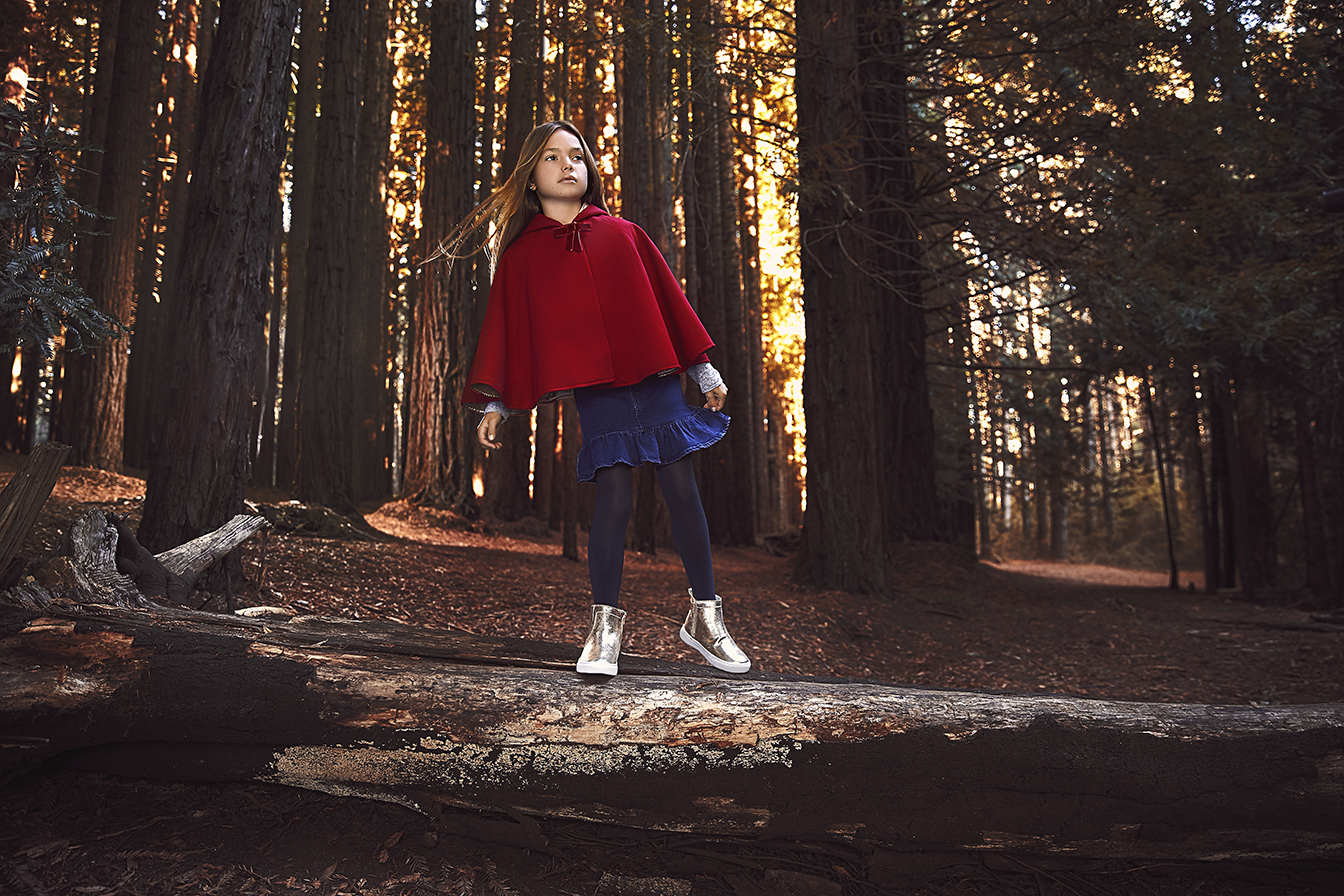 A young girl in a red cape stands on a fallen log in a sunlit forest wearing a blue dress and gold boots for a fairytale fashion look.
