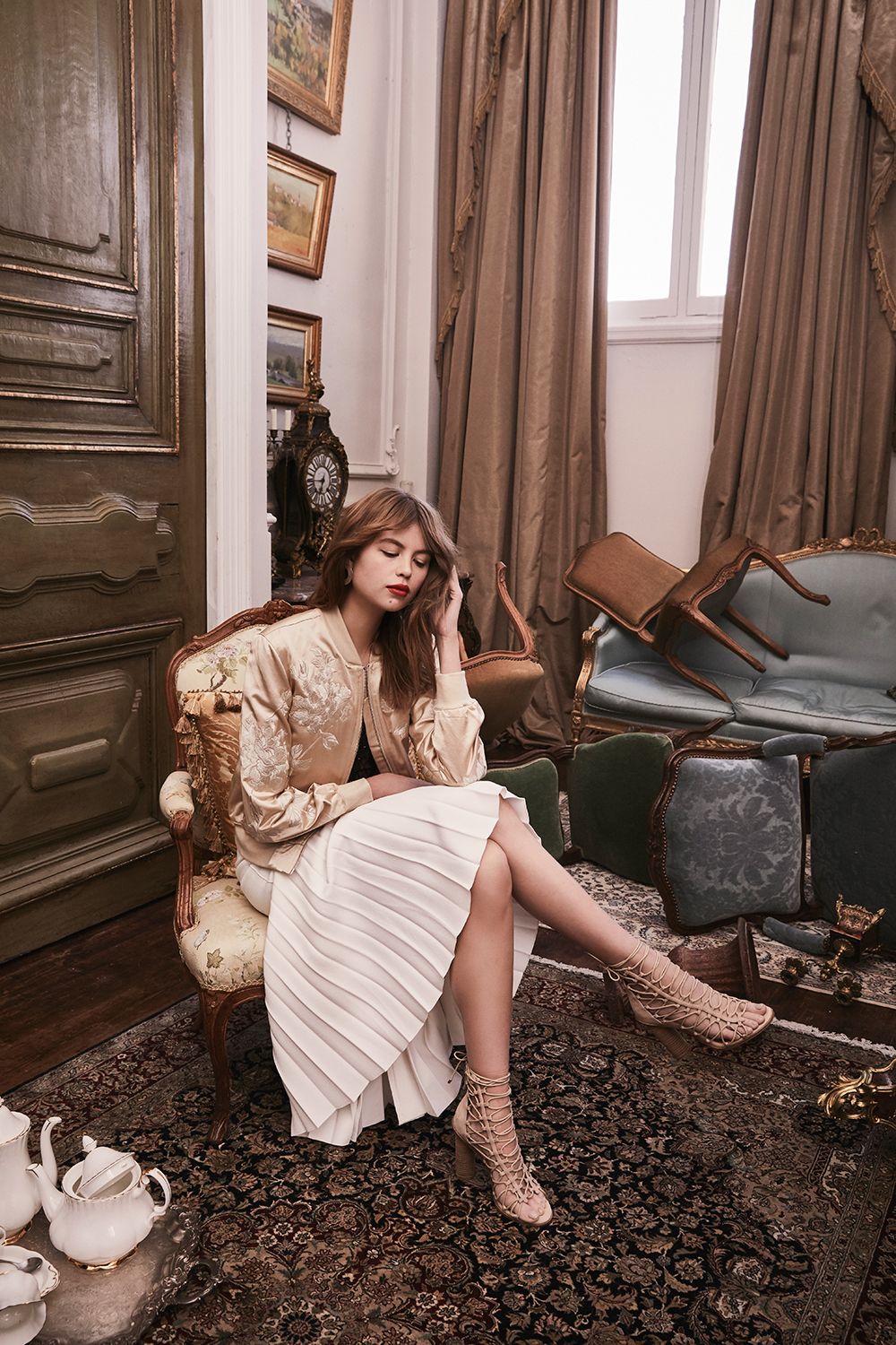 An adult woman seated in a vintage chair wearing a gold jacket and pleated skirt, indoors with antique furnishings and decor.