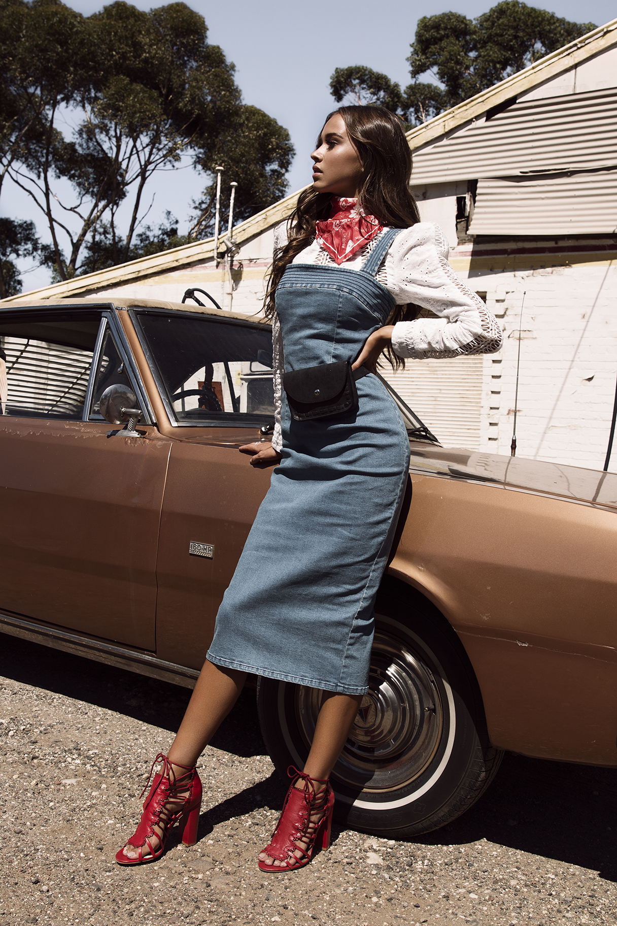 A woman in a denim dress leans against a bronze car with a bandana and red heels in front of a corrugated metal building.
