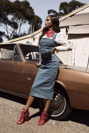 A woman in a denim dress leans against a bronze car with a bandana and red heels in front of a corrugated metal building.