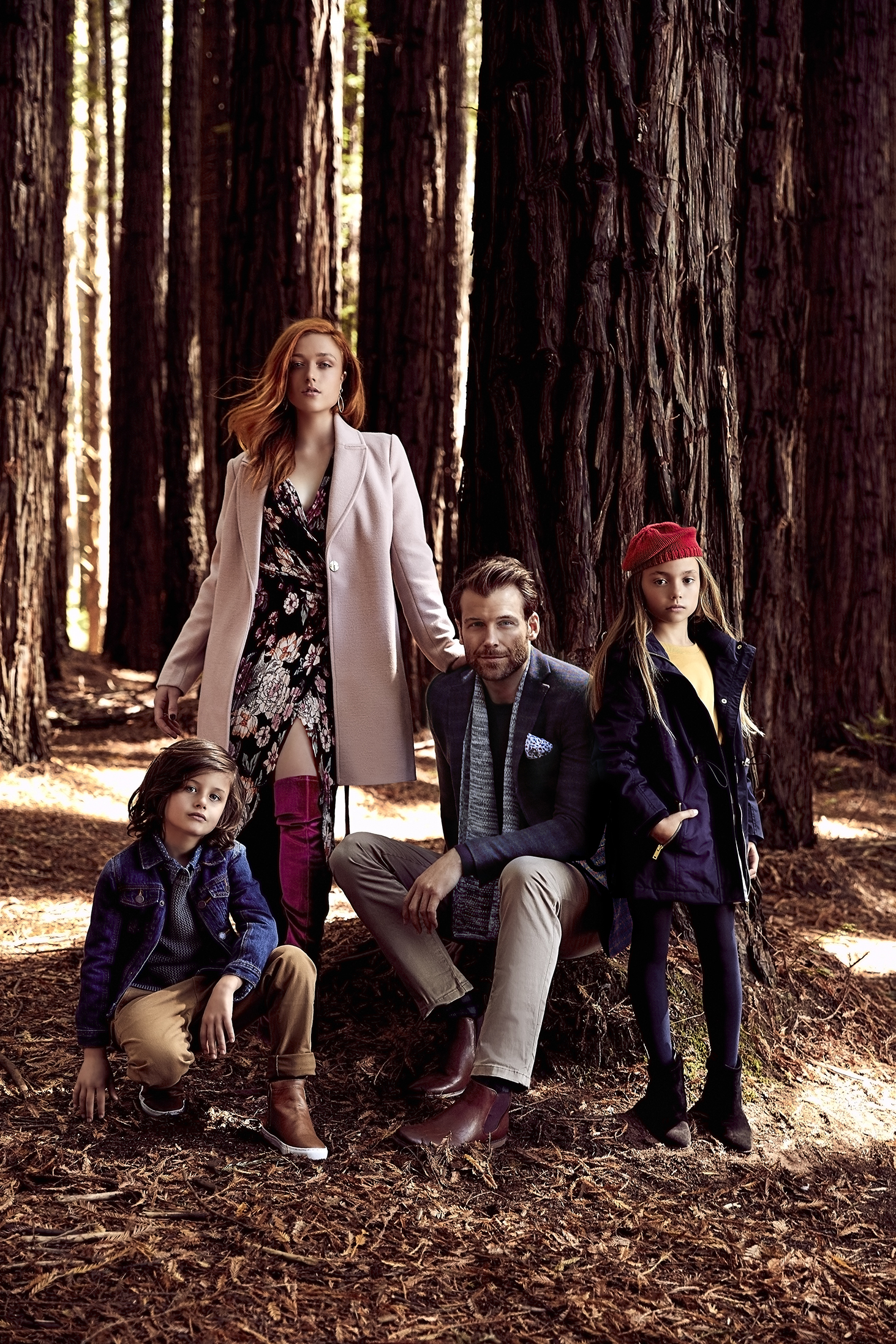 Family of four poses in a forest of tall, bare trees. Woman wears floral dress and pink coat; man in blazer; children in jackets.
