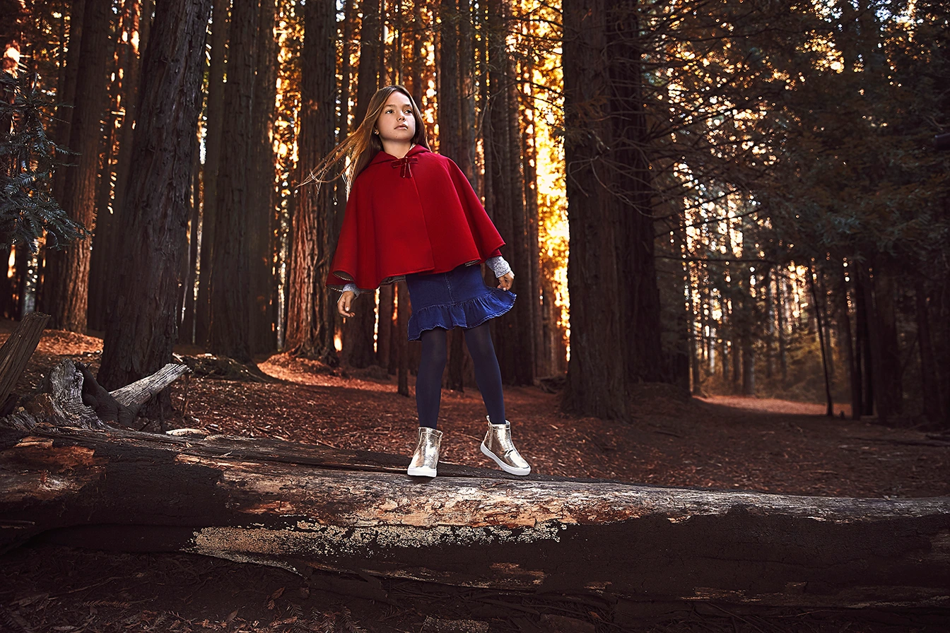 Young girl in a red hooded cape and blue skirt standing on a large fallen log in a dense redwood forest.