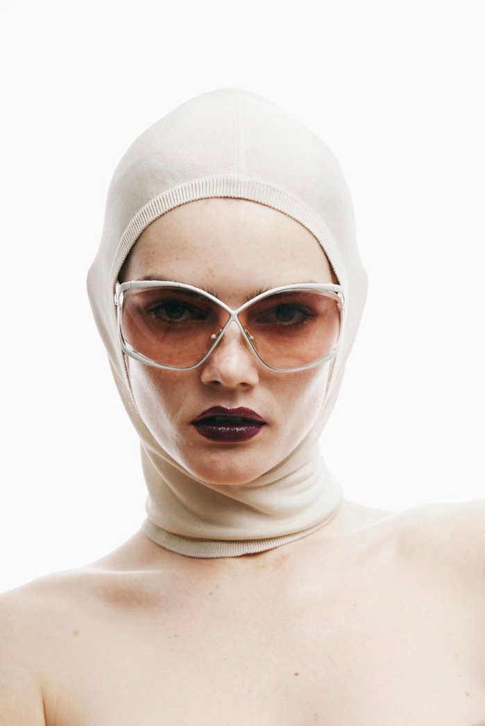 Close-up portrait of a model with pale skin, wearing a head covering and oversized sunglasses against a stark white background.