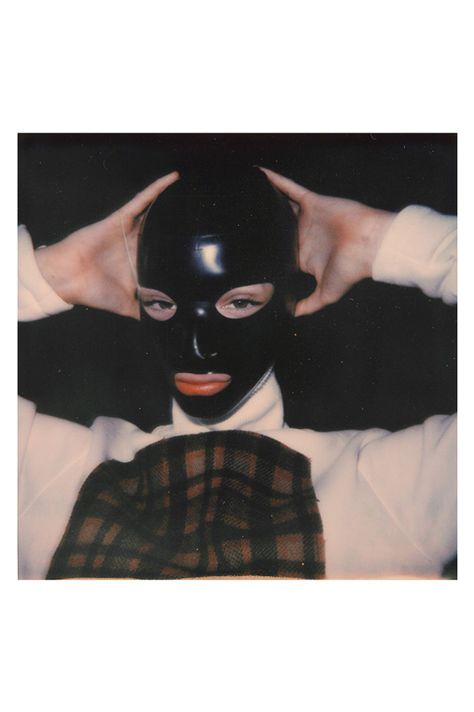 Person wearing a black latex mask, white turtleneck, and plaid garment against a dark background, with their hands behind their head.