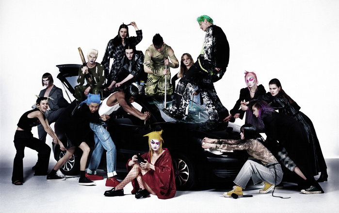 A diverse group of adults surrounds a black car against a white backdrop, creating a fashion editorial image with dynamic posing and expressions.