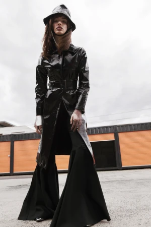 Fashion-forward woman in a black patent trench coat and wide-leg pants standing outdoors against an industrial backdrop and cloudy sky.
