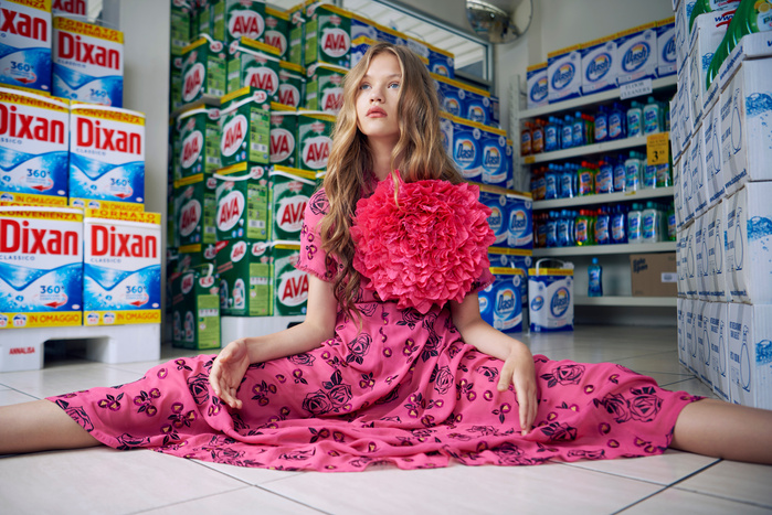 Young girl in pink dress with flower detail sitting cross-legged in the aisle of a store surrounded by cleaning products on shelves.