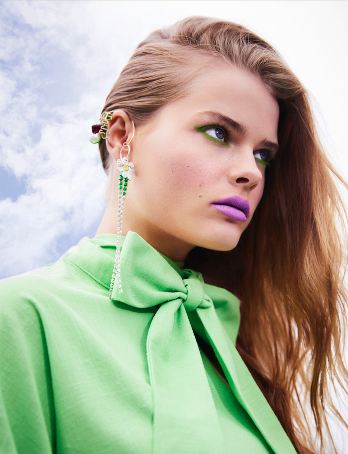 Close-up portrait of a young woman with colorful eye shadow and lipstick, wearing a light green blouse and statement earrings against cloudy sky.