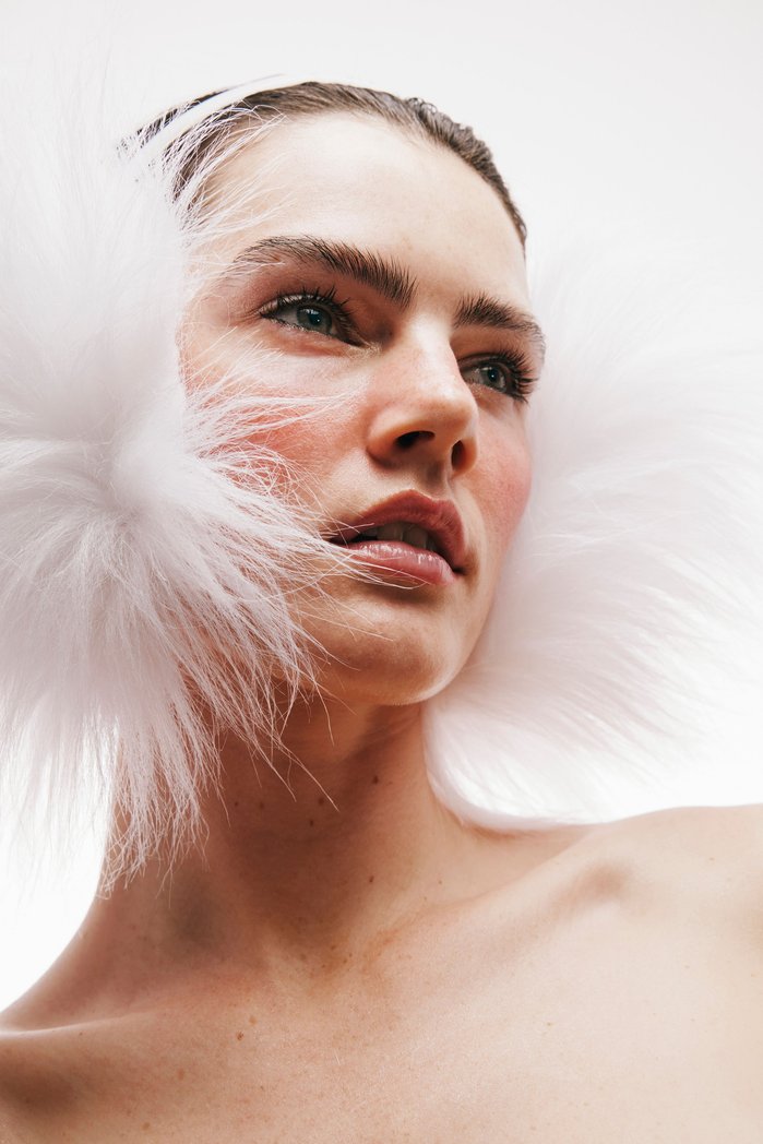 Portrait of a young fair-skinned woman gazing upwards, adorned with fluffy white accents around her head against a stark white background.