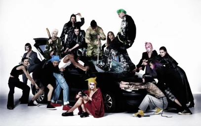 Group of models in avant-garde outfits posing around a wrecked black car on a white background, styled by Brittni Morrison