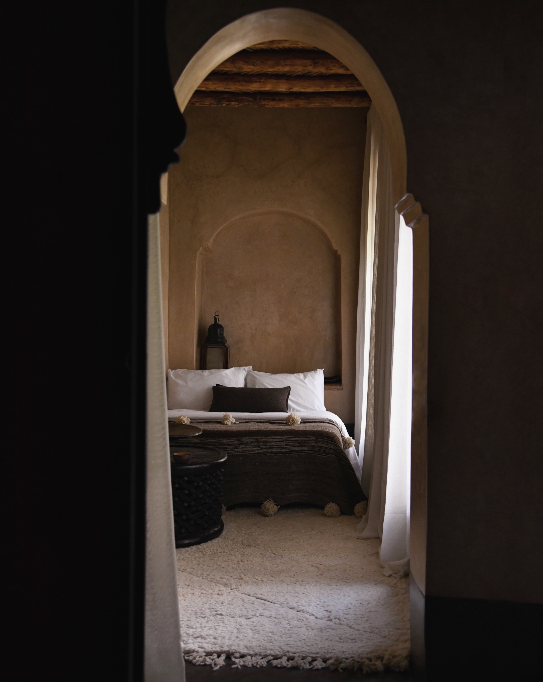 Bedroom interior with a bed, arched doorway, white rug, carved wood table, and neutral colour palette creating a serene, elegant space.